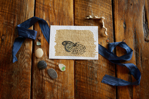 Found butterfly wing (Monarch/ Lepideroptera / Danaus plexippus), ink, acrylic paint, and gold leaf on 5x7 cold press watercolor paper, shown on aged oak background with blue silk ribbon, driftwood, sea glass and pebbles. 